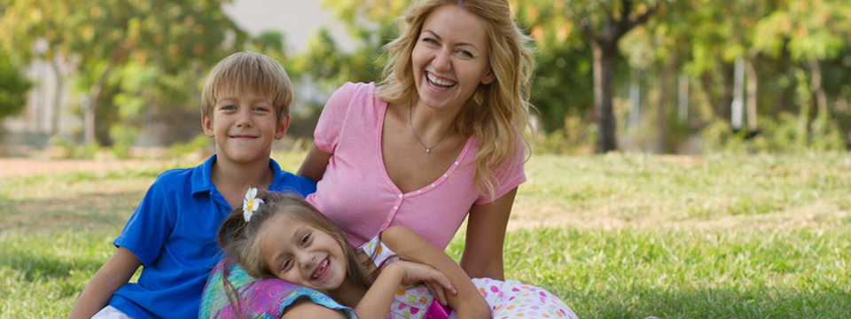 Photo of a mom with her son and daughter sitting on the grass in a park on a beautiful sunny day smiling and laughing into the camera.