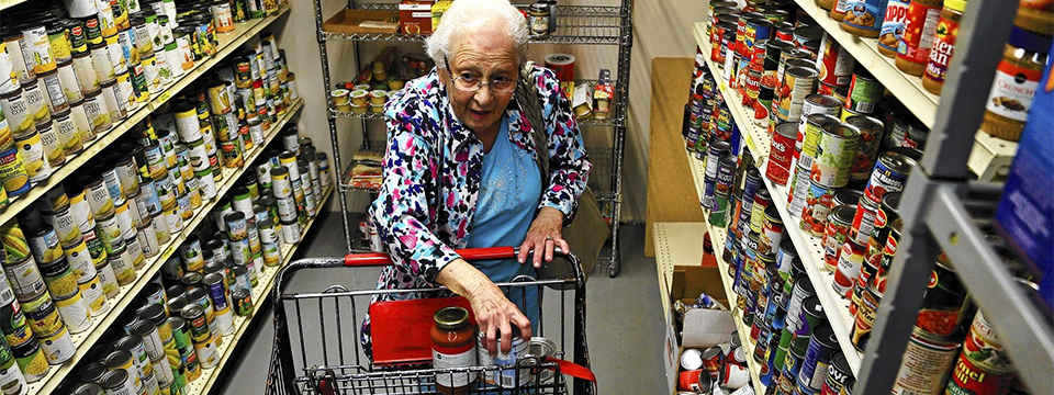 Photo of an elderly woman standing alone putting things into her shopping cart in a grocery store aisle.