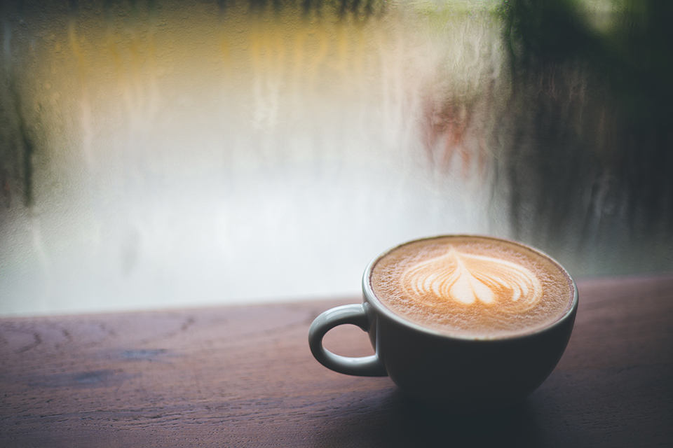 A beautifully frothed coffee latte sits atop a wooden table facing a window streaked from a light rain outside.