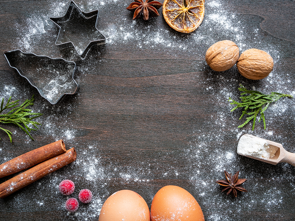 On top of a wooden table covered in sprinkles of white flour are cinnamon sticks, pine fronds, raspberries, star anise, eggs and cookie cutters all arranged in a circle.