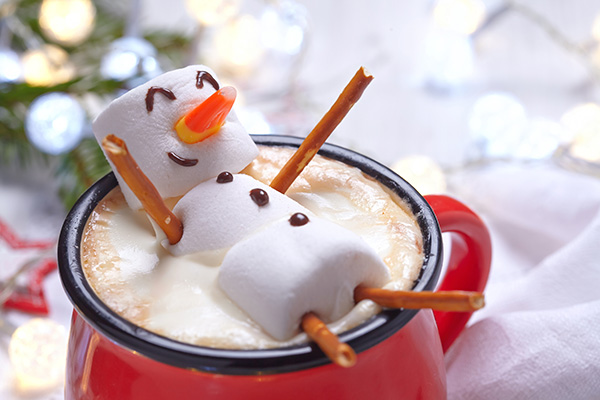 On top of a red mug filled with hot chocolate floats a smiling marshmallow snowman.
