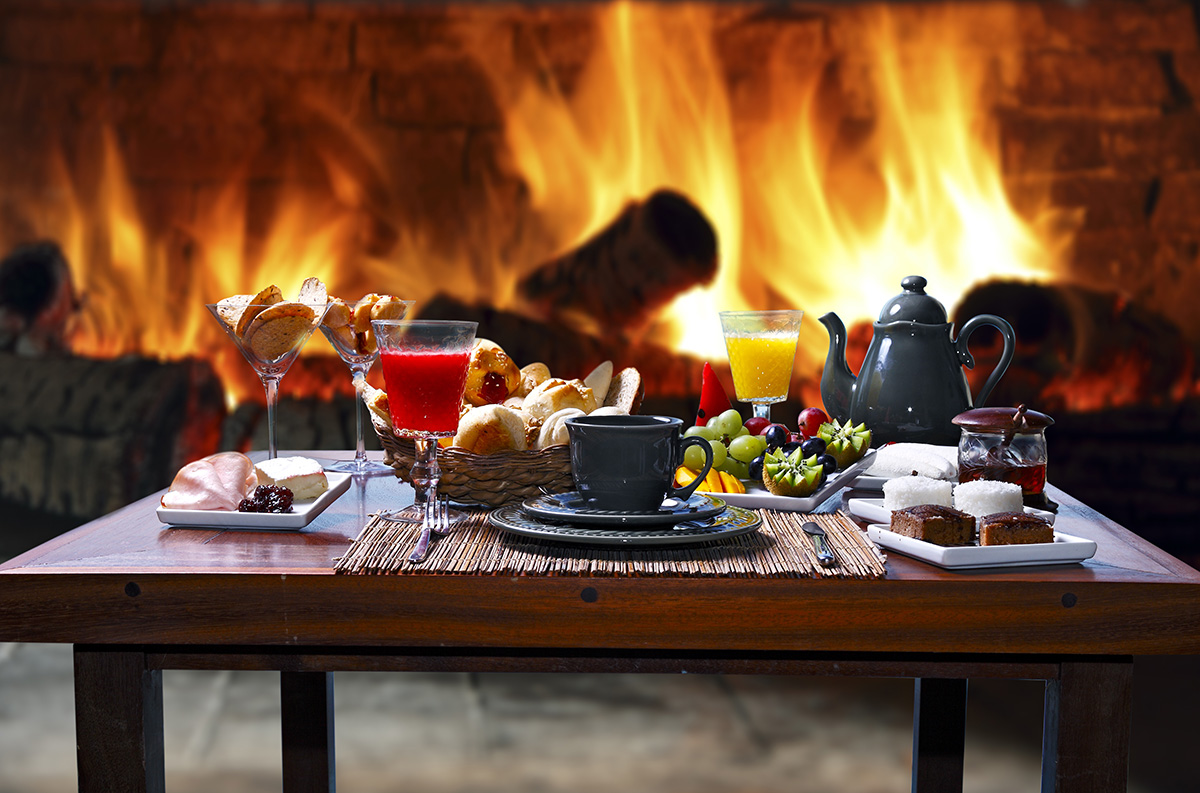 A beautiful breakfast of fruit, bread, pastry, juice and coffee sit atop a wooden table placed before a roaring fireplace.
