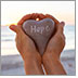 Hands hold up a heart-shaped stone with the word Hope inscribed on it. Ocean in the background.