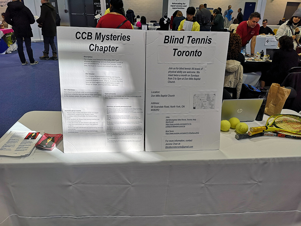 Side by side table placards. On the left the CCB Mysteries Chapter placard. On the right the Blind Tennis Toronto placard. Expo in the background.