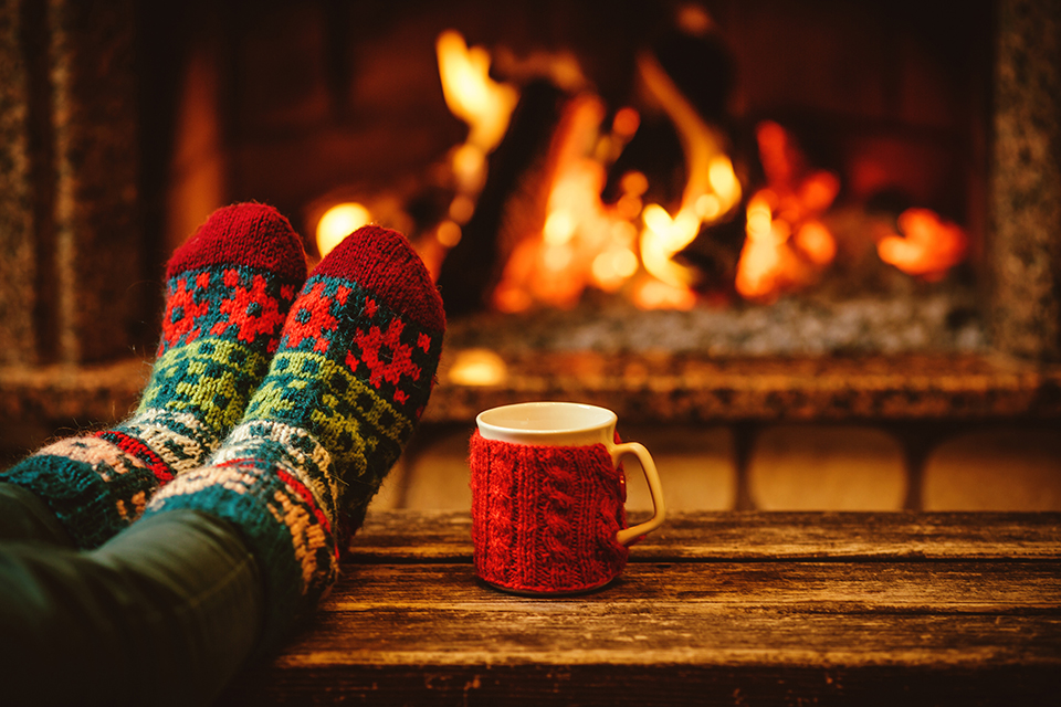 A woman in festive holiday socks has her feet up on a wooden table. Hot chocolate with knitted warmer on cup. Fireplace in background.
