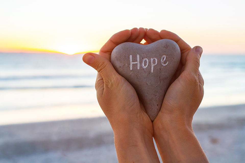 Hands hold up a heart-shaped stone with the word Hope inscribed on it. Ocean in the background.