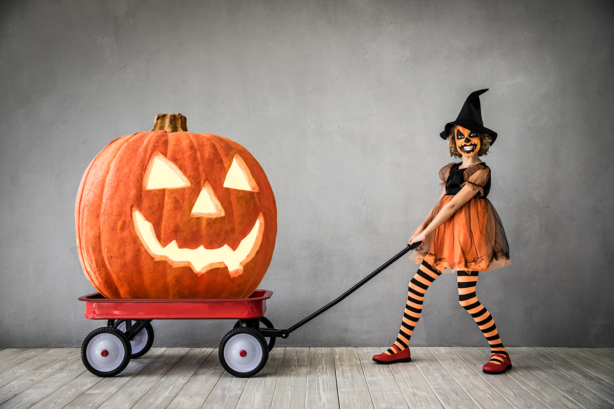 A little girl dressed up as a pumpkin witch pulls a red wagon with a huge jack-o-lantern inside it.