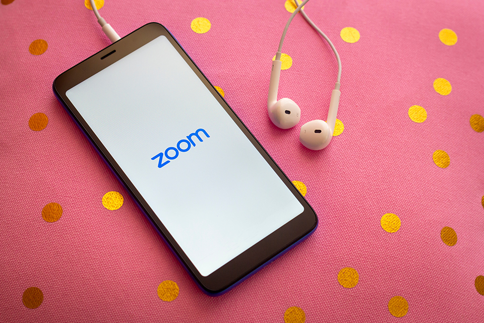 On a pink table cloth with gold dots sits a smart phone with the Zoom app open and a pair of wired ear buds connected to the phone.