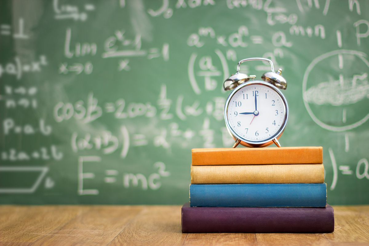 On a table sits a stack of 4 nondescript books with an alarm clock on top showing a time of 9 o'clock. Green chalkboard in the background filled with equations.
