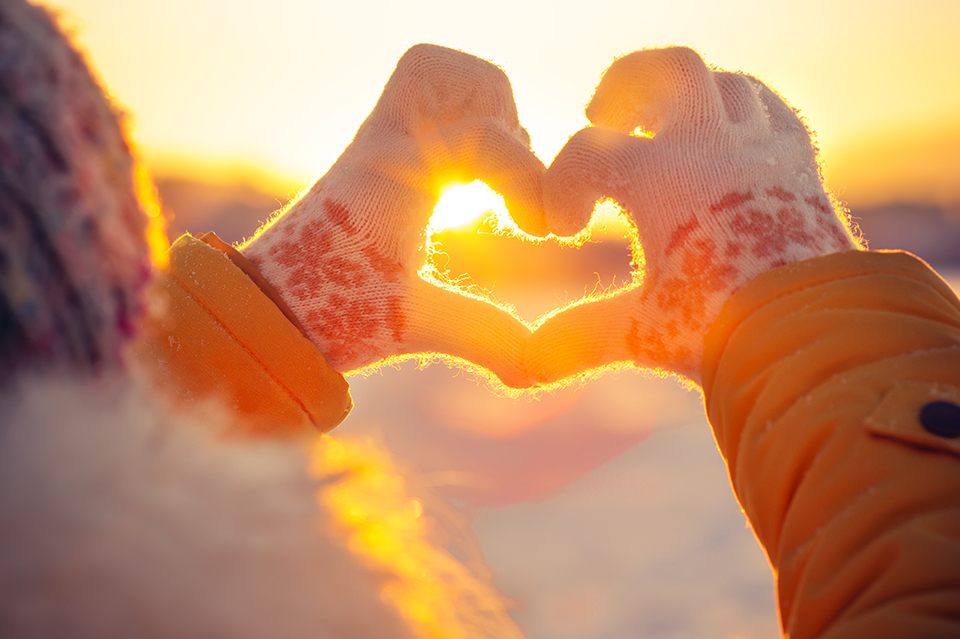 A woman's hands in winter gloves makes a heart symbol through which shines a sunset on a cold winter's day.
