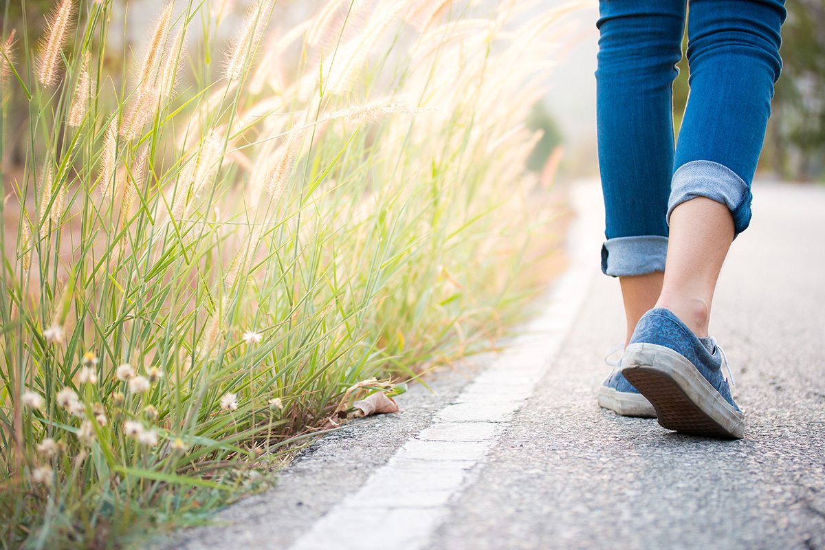 A woman in jeans and sneakers walks along a roadside covered in grass, brush and flowers.