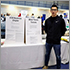Jerome Chan stands to the right of side by side table placards. On the left the CCB Mysteries Chapter placard. On the right the Blind Tennis Toronto placard. Expo in the background.