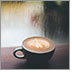 A beautifully frothed coffee latte sits atop a wooden table facing a window streaked from a light rain outside.