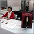 Photo of Donna Jodhan sitting at a table at the Expo 2018. On the table in front of her are fliers, cards, pamphlets, as well as a stand up sign announcing her Dinner Mystery Evenings.